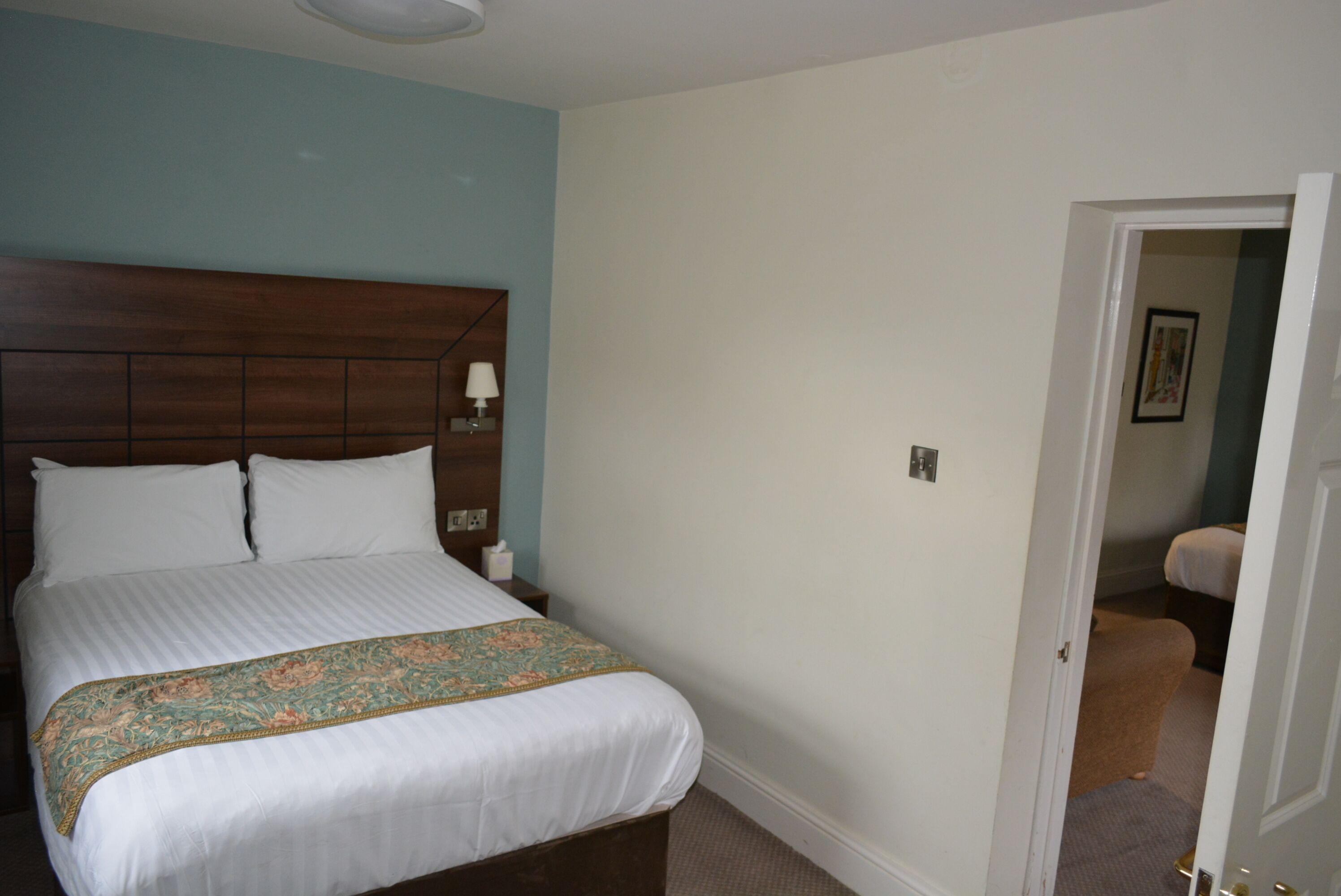 Un dormitorio con cabecera de madera, cama con ropa blanca y un plaid floral, mesita de noche con lámpara, y una puerta que da a otra habitación.