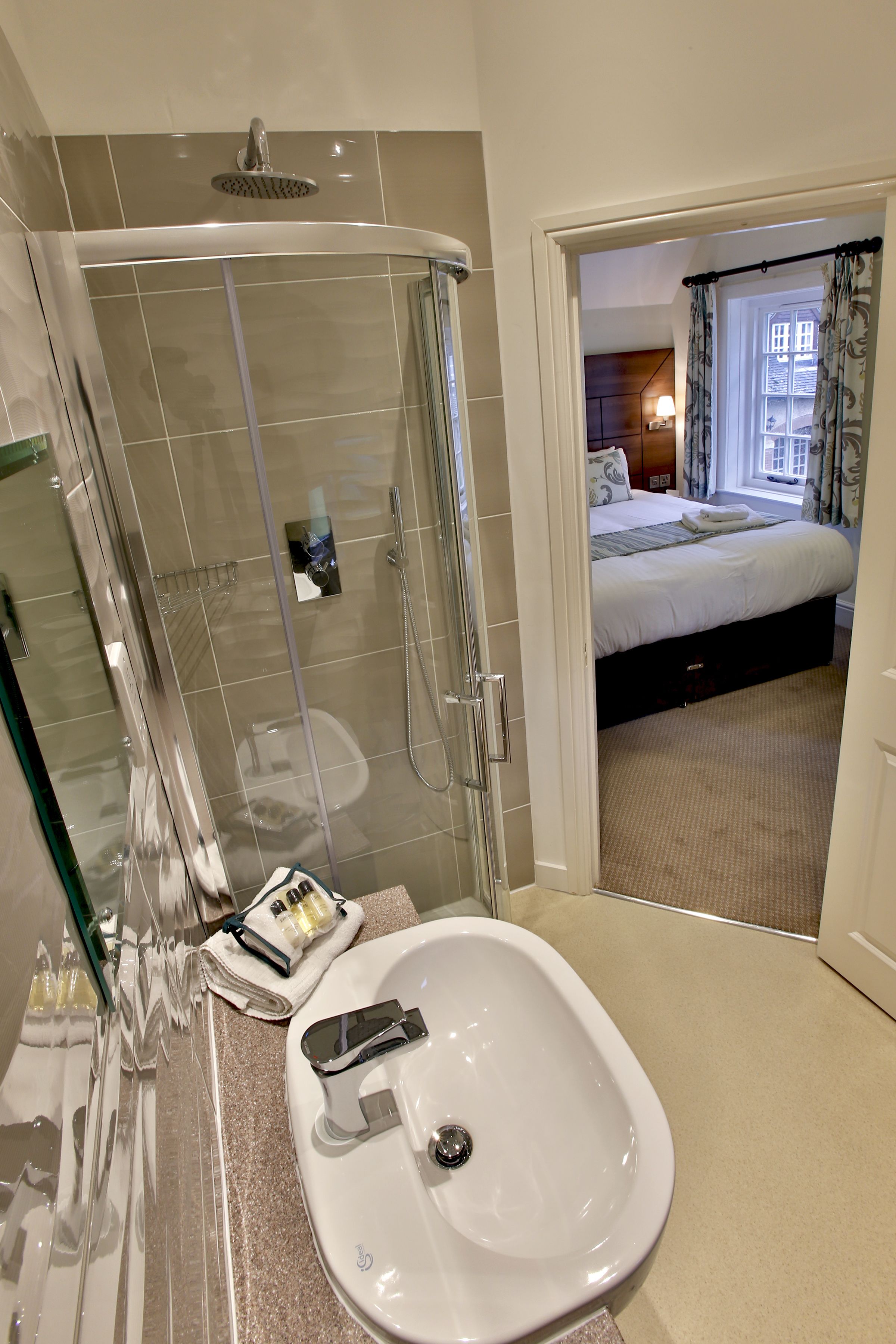 A hotel bathroom with a shower, sink, and a view of a bedroom.