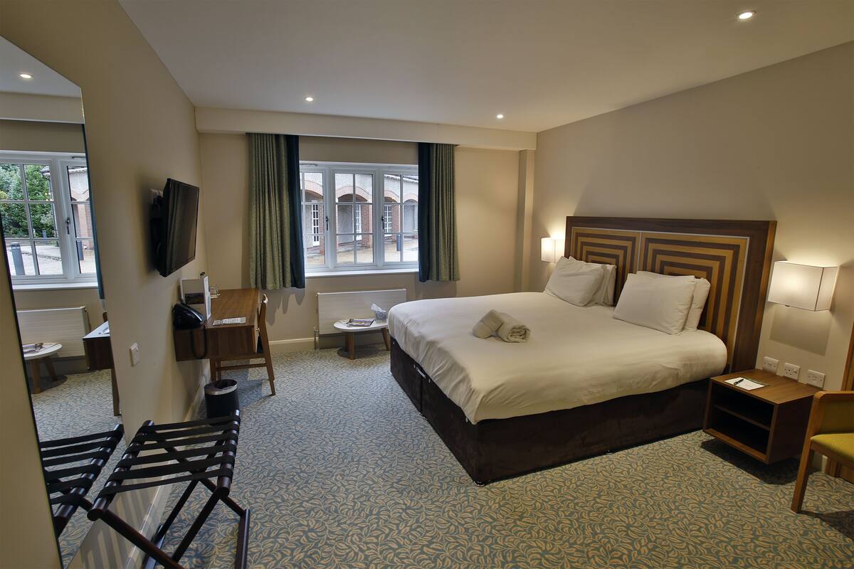 A hotel room with a large bed, a desk with a chair, a TV mounted on the wall, and a window with curtains.