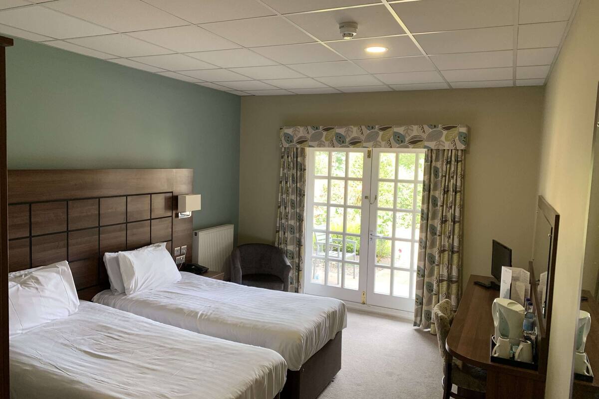 A hotel room with two single beds, a wooden headboard, a desk with a chair, a radiator, and a window with curtains.
