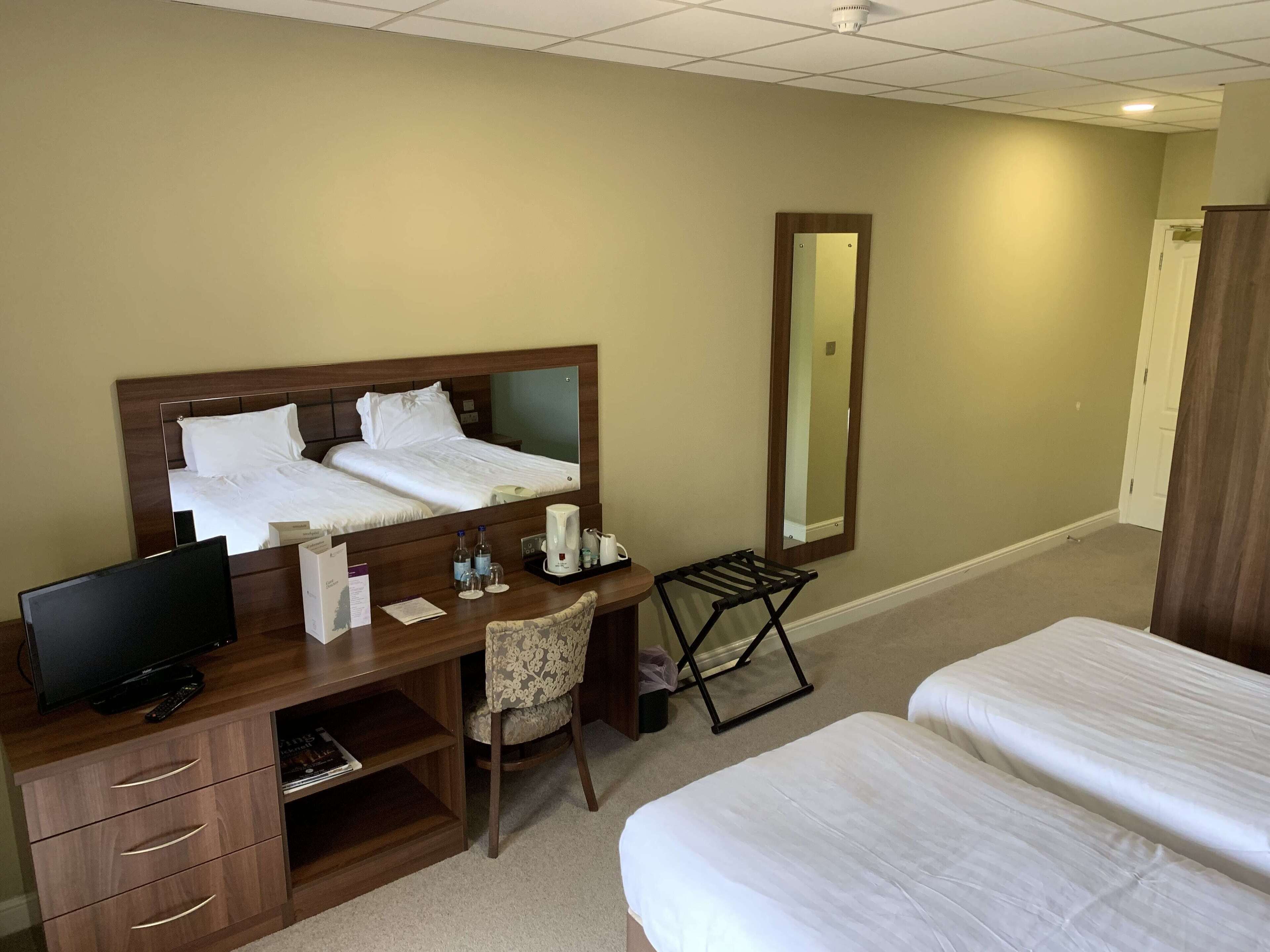 A hotel room with a bed, a desk with a computer, a chair, a coffee maker, and a mirror.