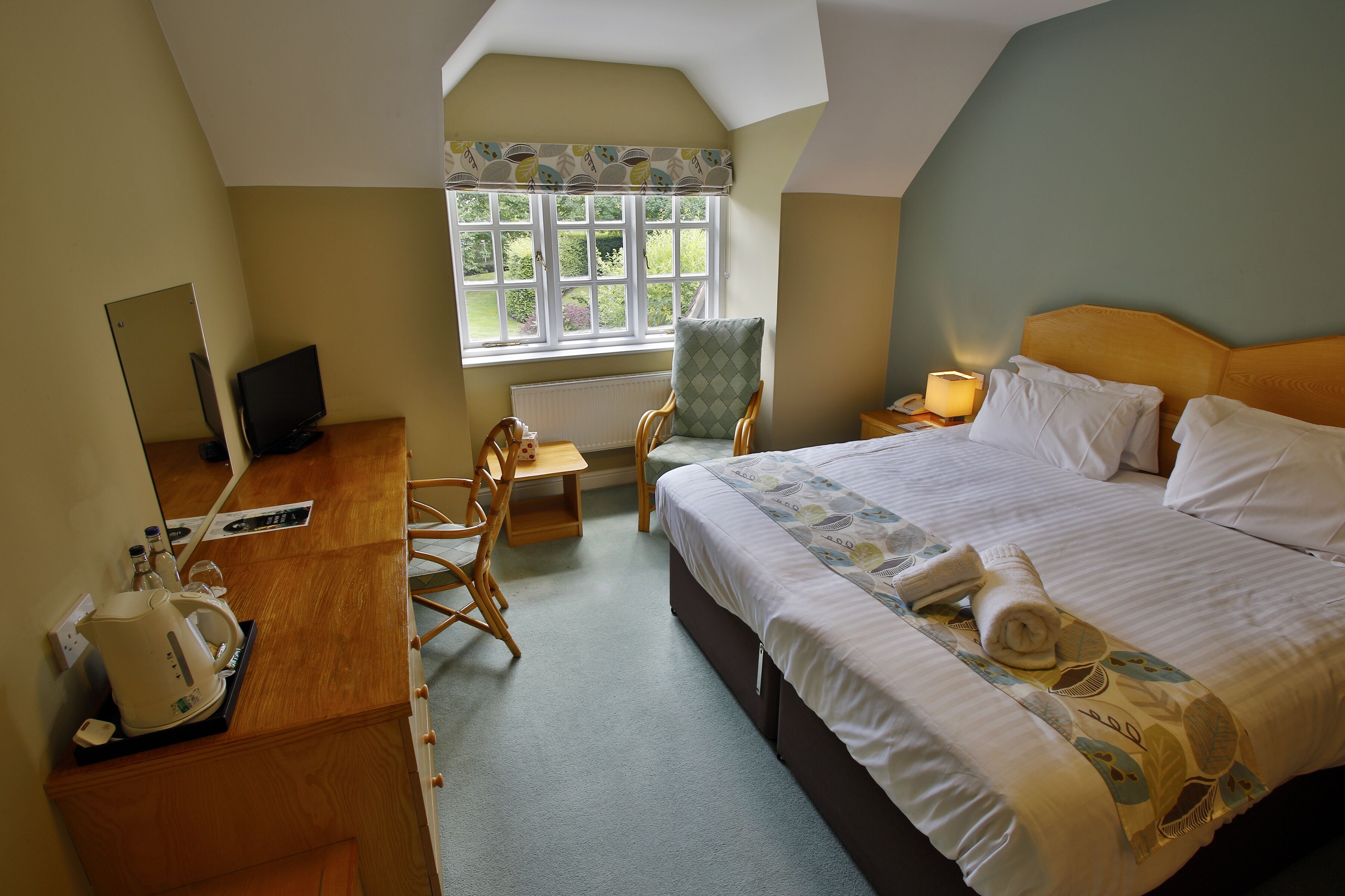 A hotel room with a bed, a desk with a computer, a chair, a window with a view, and a nightstand with a lamp.