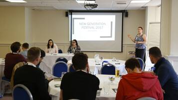 A conference room with a presentation screen displaying "NEW GENERATION FESTIVAL 2017" at Quinn Grand Hotel. Attendees are seated at round tables with nameplates, and a speaker is addressing the audience.