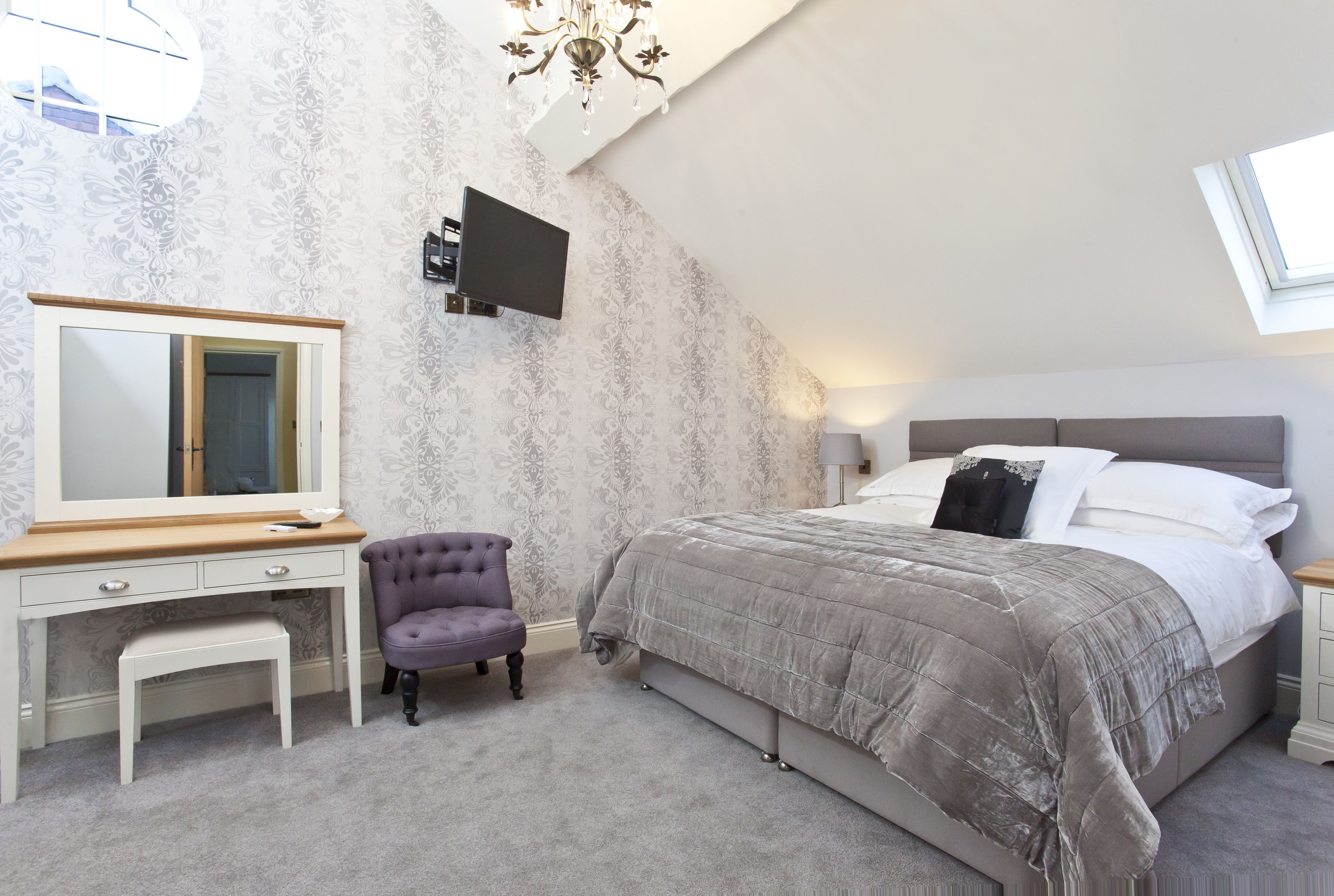 A bedroom with a bed, a vanity with a mirror, a chair, and a television mounted on the wall.