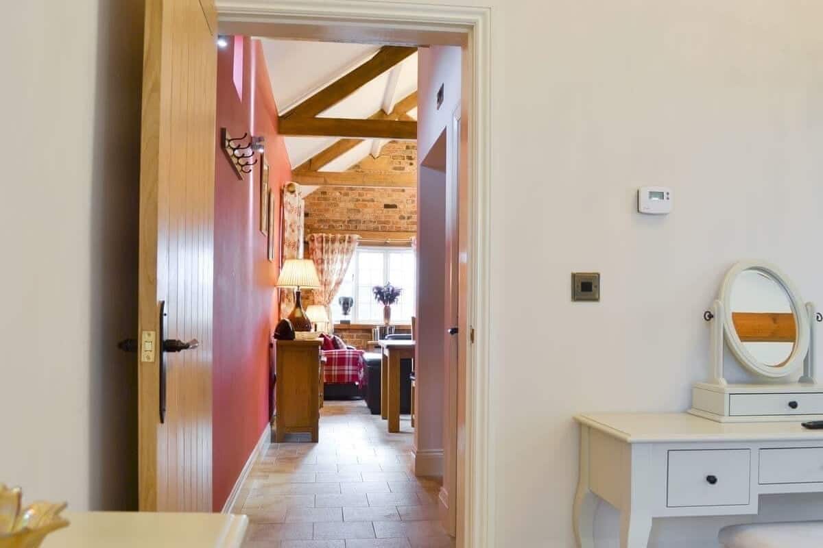 A narrow hallway leading to a dining area with a brick wall and wooden beams. A white vanity with a mirror and a small table with a lamp is visible.