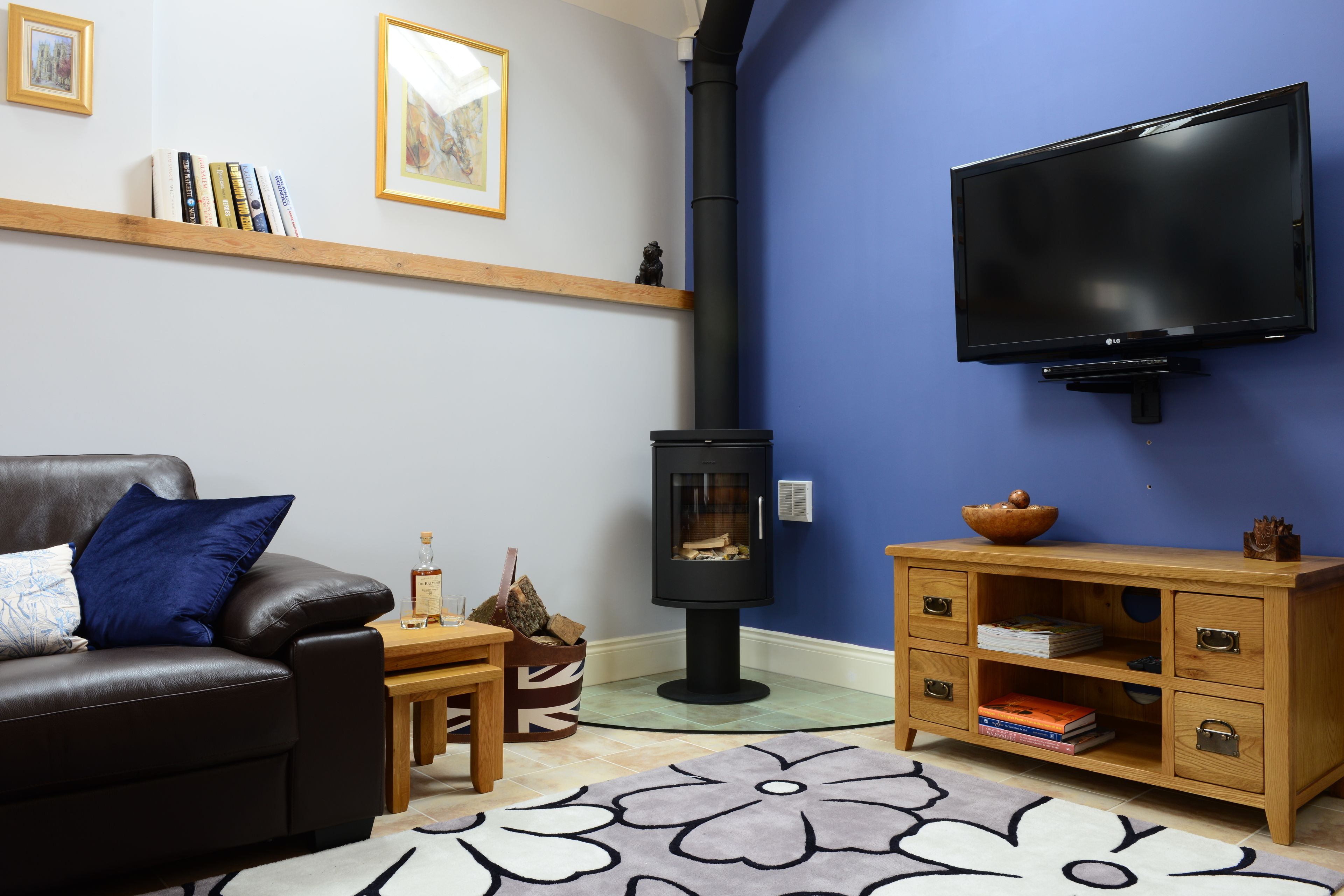 A living room with a black leather sofa, a wooden coffee table, a fireplace, a TV mounted on the wall, and a wooden cabinet with drawers.