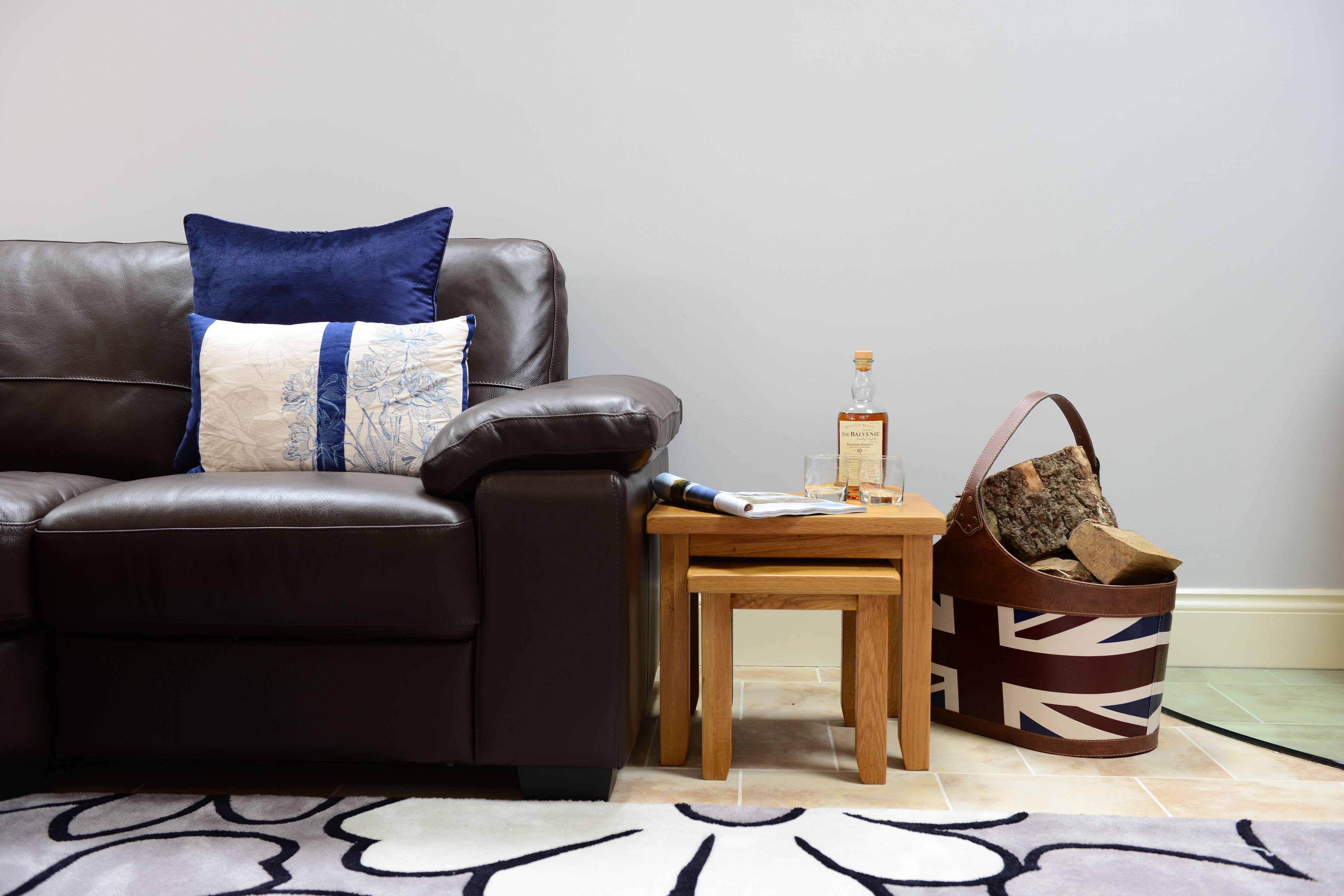 A brown leather sofa with a blue and white pillow, a small wooden table with a bottle and papers, and a brown bag with a Union Jack pattern.