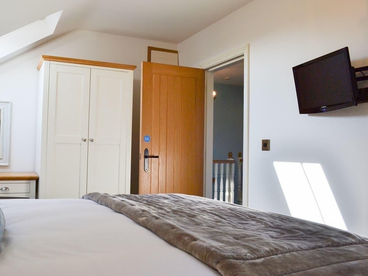 A bedroom with a bed, a wooden door, a television mounted on the wall, and a window providing natural light.