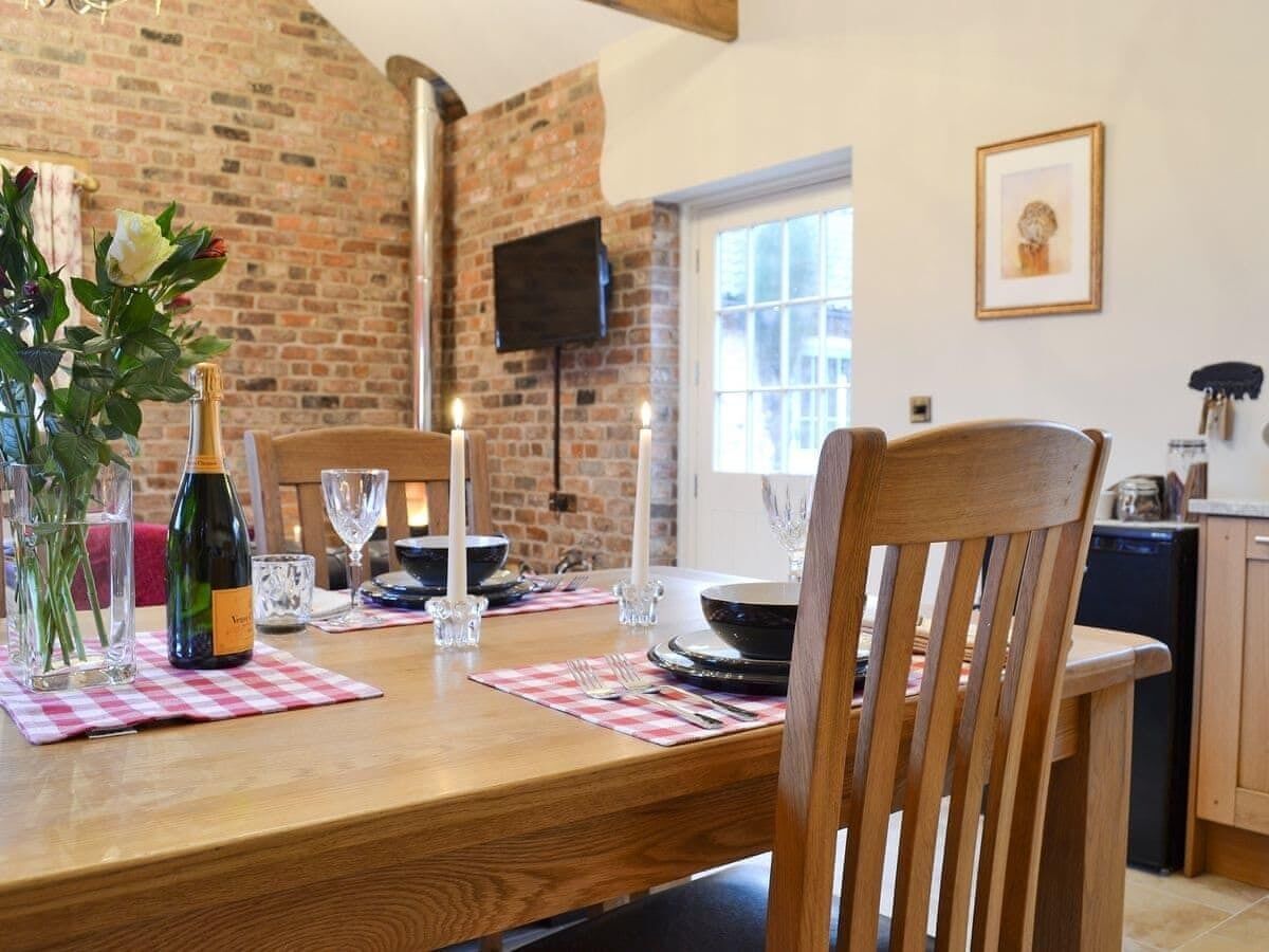 Un área de comedor con una mesa de madera preparada para dos personas, una botella de vino y velas. Se aprecia una pared de ladrillo y un televisor al fondo.