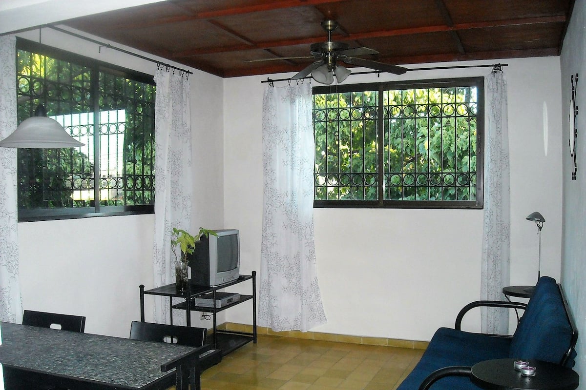 A room with a dining table, chairs, a blue sofa, a TV on a stand, and a ceiling fan.