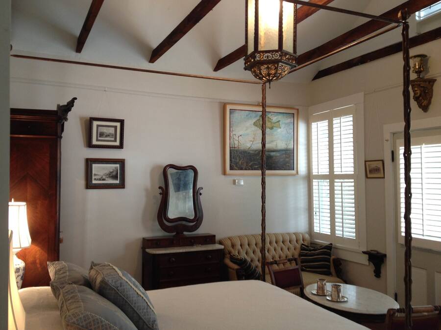 A room with a bed, a wooden chair, a table with a cup, a mirror, and a painting on the wall.