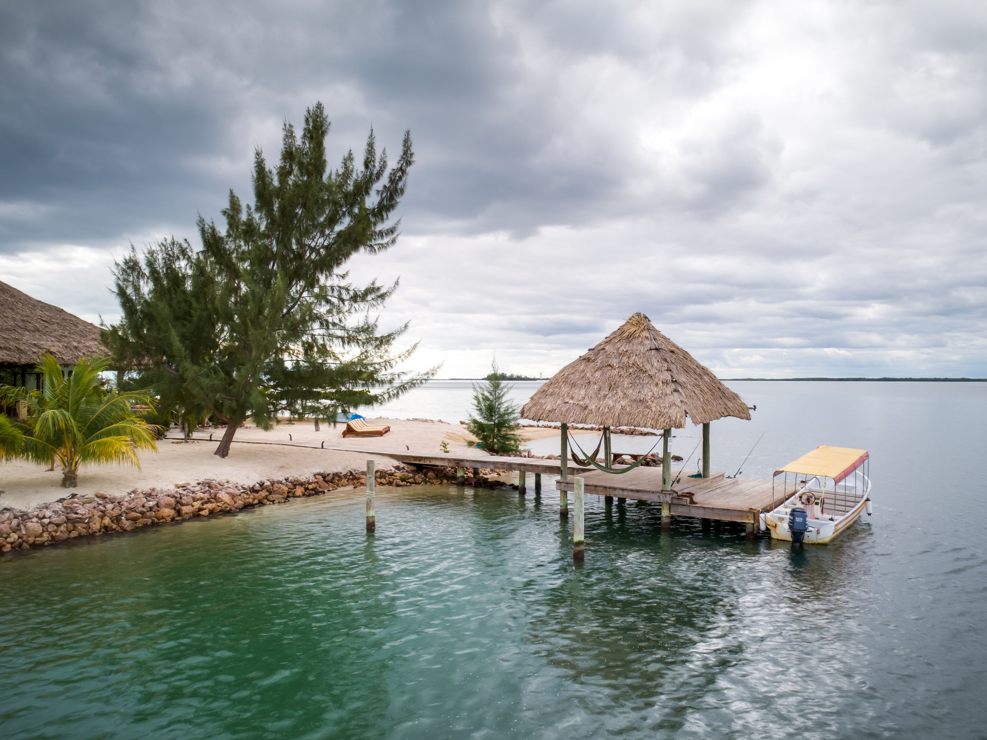 A tranquil beachside resort with a thatched-roof hut, a wooden dock extending into calm waters, and a palm tree.