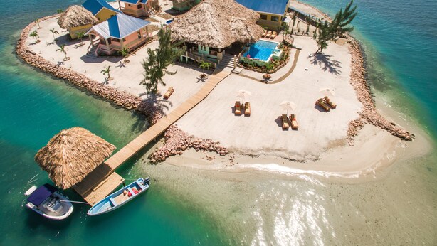 Vue aérienne d’un complexe hôtelier tropical situé sur une île, avec des bâtiments aux toits de chaume, une piscine et une jetée où sont amarrés des bateaux.