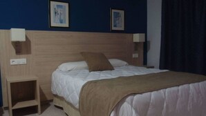 A neatly made bed with a white duvet and a brown pillow, a wooden nightstand, and two wall-mounted lamps. - Catalan Hotel (Puerto Real)
