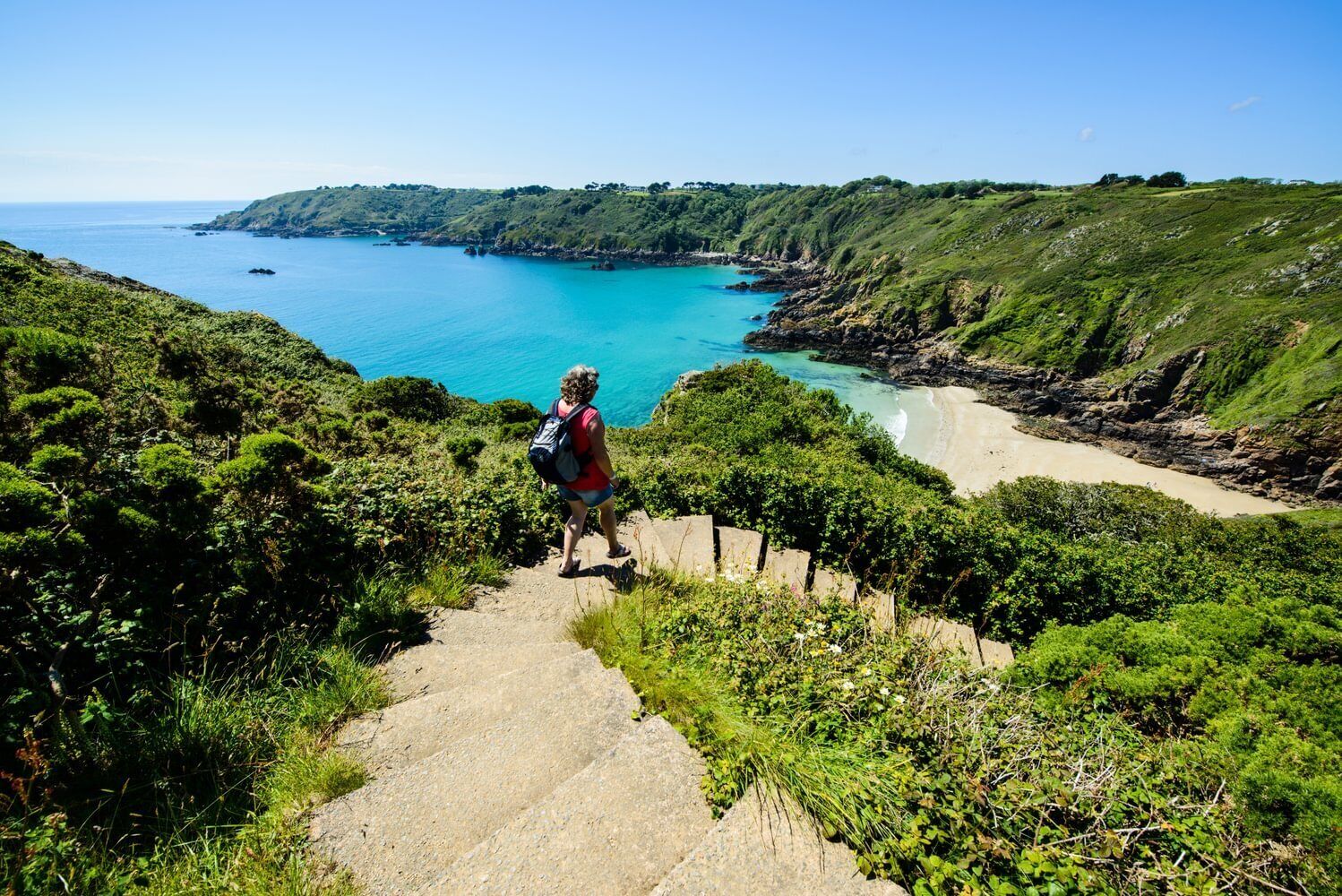 A person hiking on a stone path with a backpack, surrounded by lush greenery and overlooking a coastal area with clear blue waters.