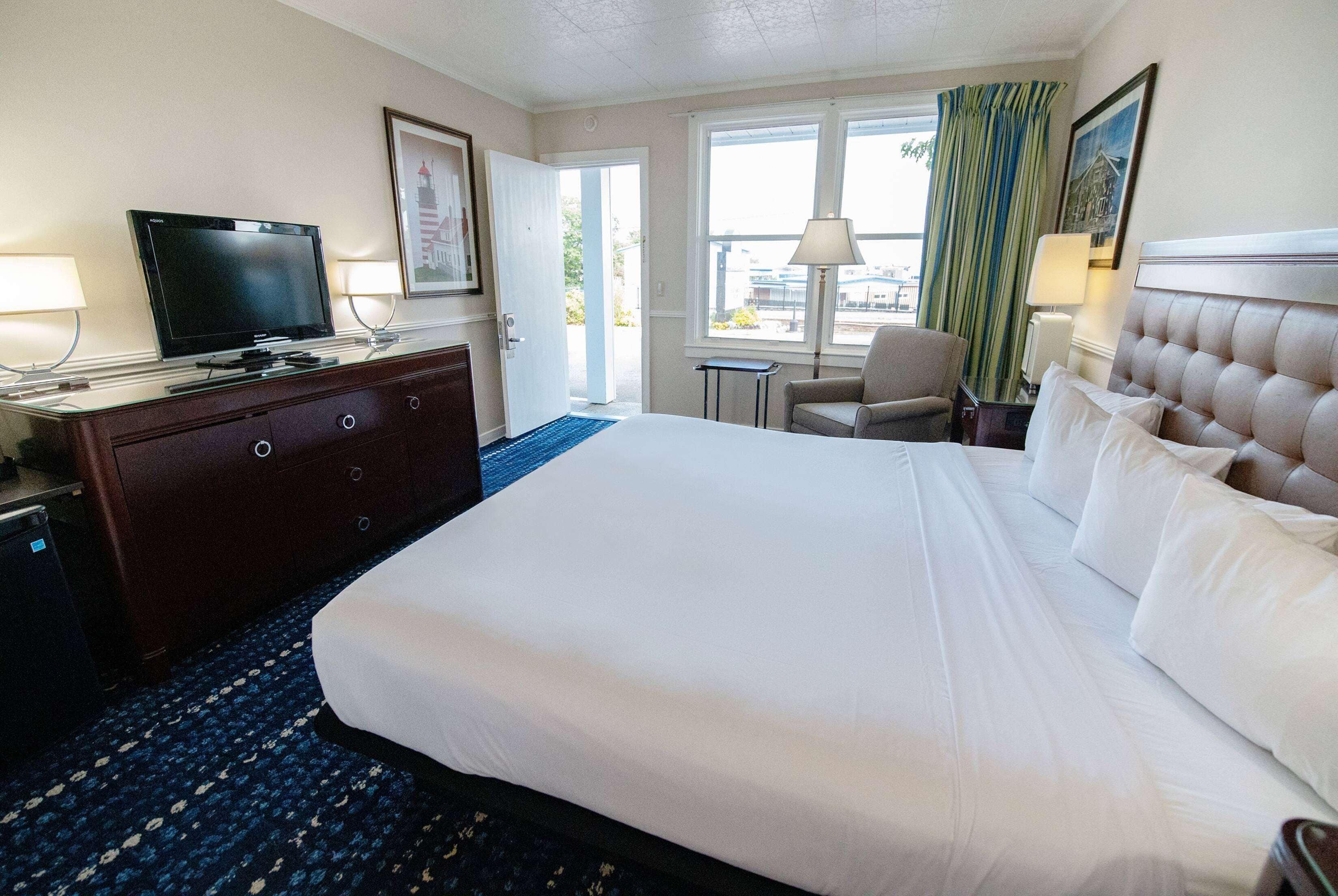 Habitación de hotel con una cama grande, un televisor en una cómoda, una silla y una ventana con cortinas.