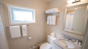 A bathroom with a toilet, sink, and a window. There are towels on a rack and a trash bin.