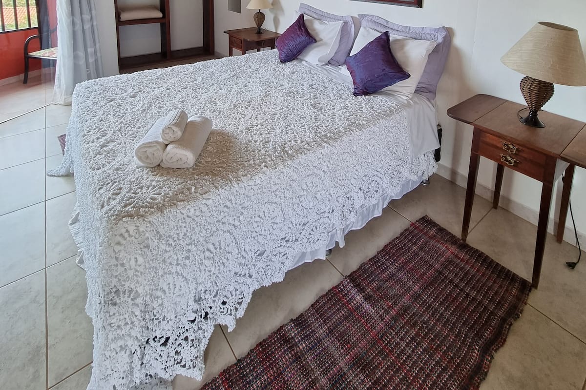 A bedroom with a large bed, a window with curtains, an air conditioning unit, a framed picture on the wall, and a small table with a lamp.
