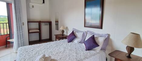 Un dormitorio con una cama grande, una ventana con cortinas, un aire acondicionado, un cuadro en la pared y una mesita con una lámpara.
