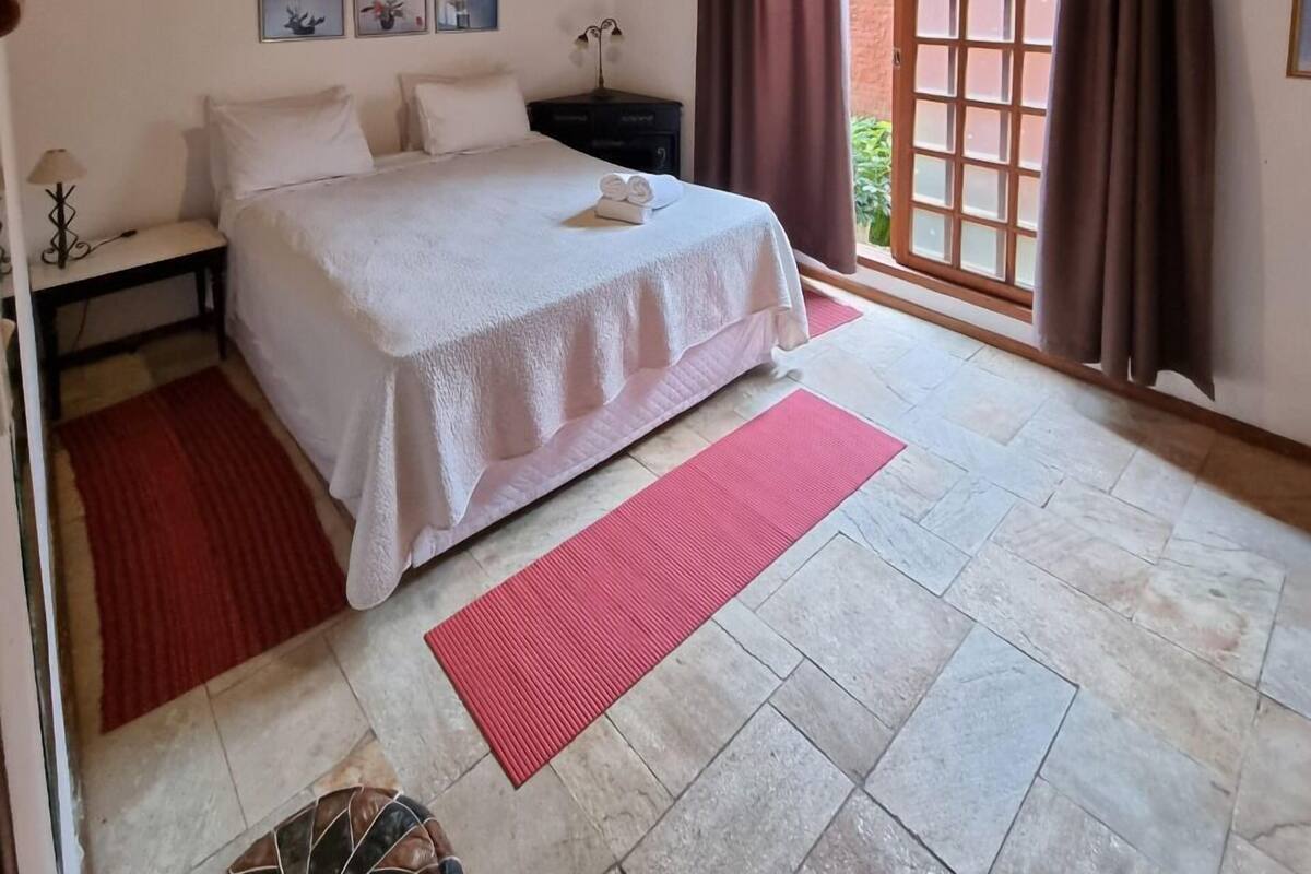 A bedroom with a wooden ceiling, a bed with white linens, a nightstand, a red rug, and a window with curtains.