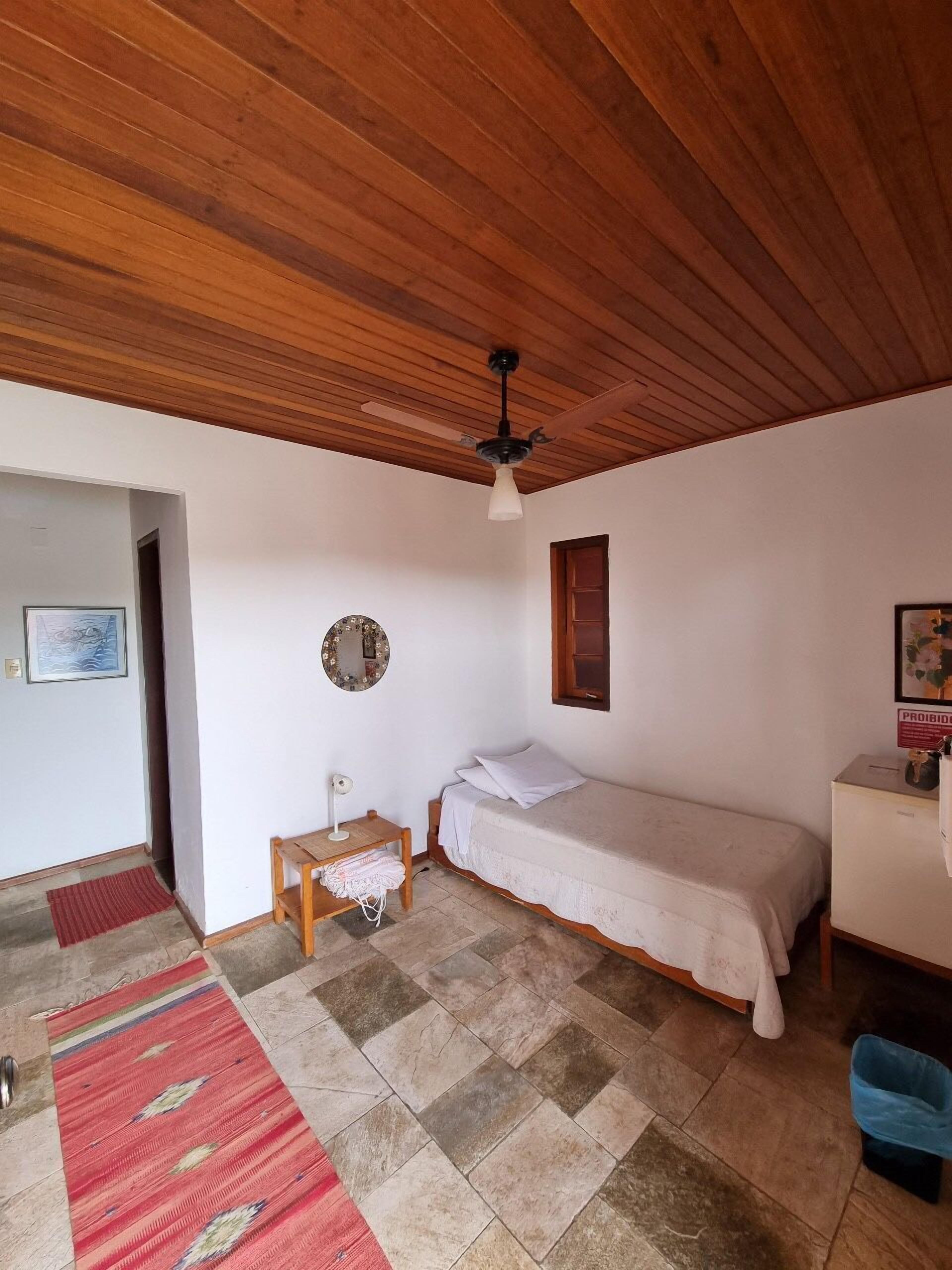 A small, single-room accommodation with a wooden ceiling, a bed, a small table, a mirror, and a red rug.