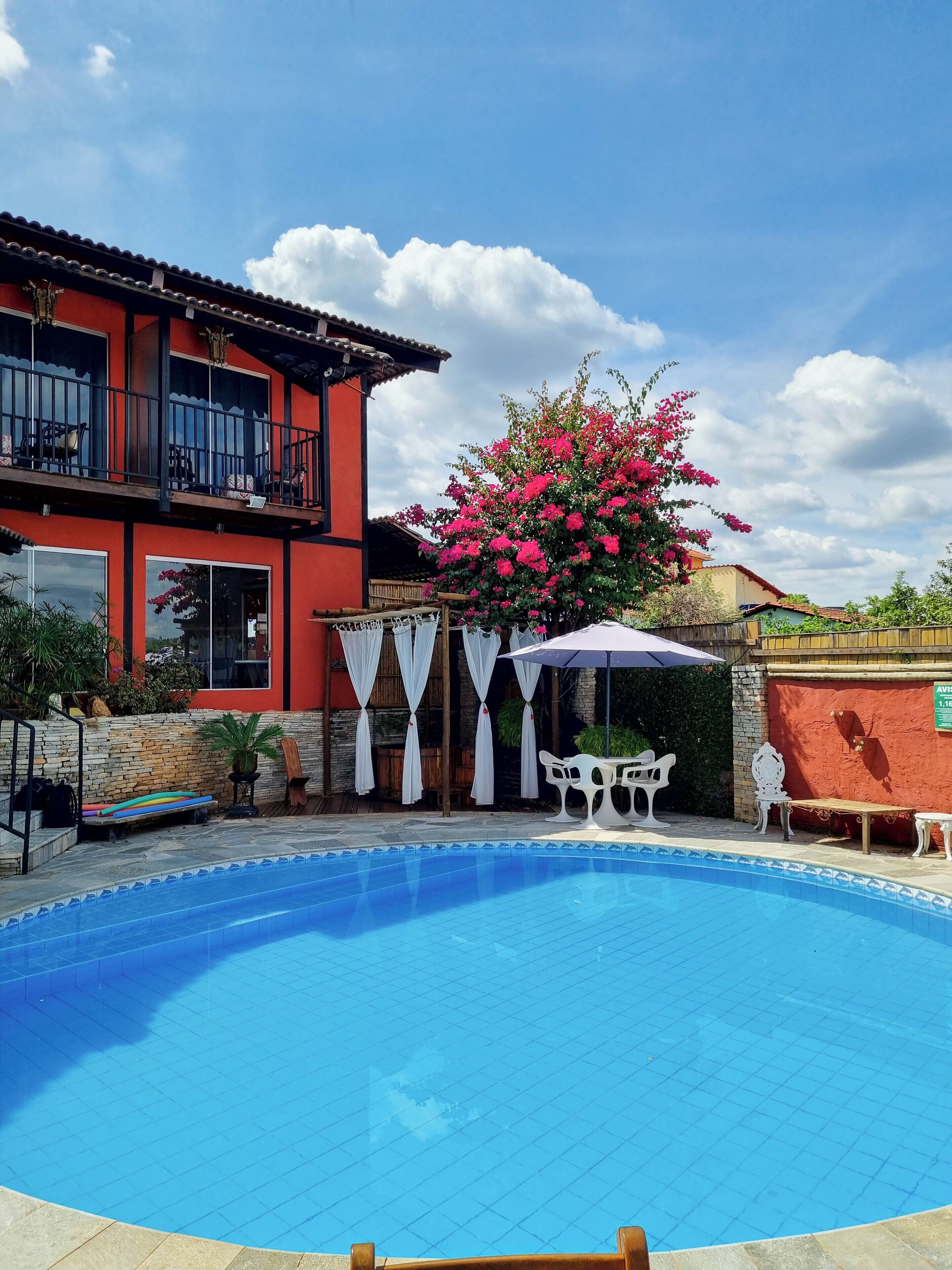A swimming pool with a tiled deck, a red building with balconies, and a large pink flowering tree.