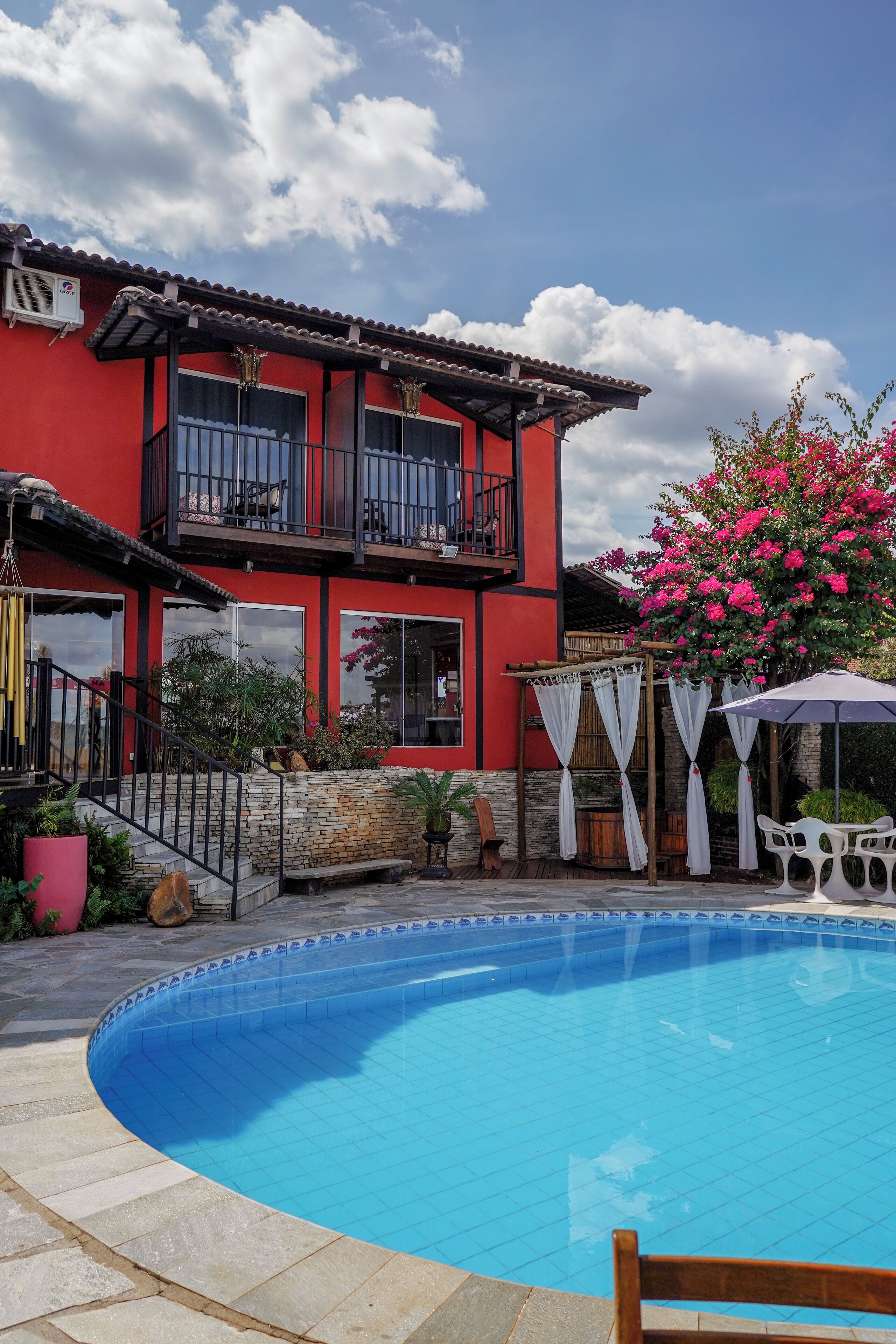 A red building with a pool, a balcony, and a pergola with white curtains.