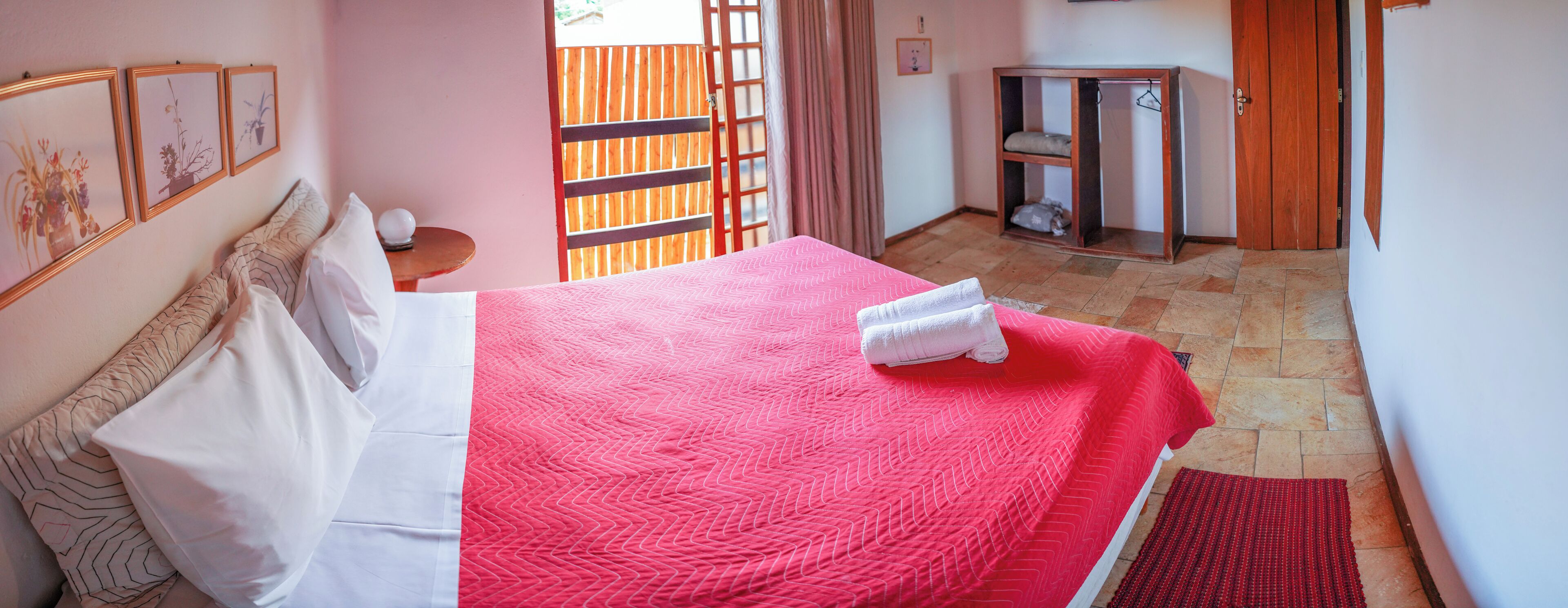 Een keurig opgemaakt bed met een rode deken, een houten nachtkastje met een lamp en uitzicht buiten door een houten deur.