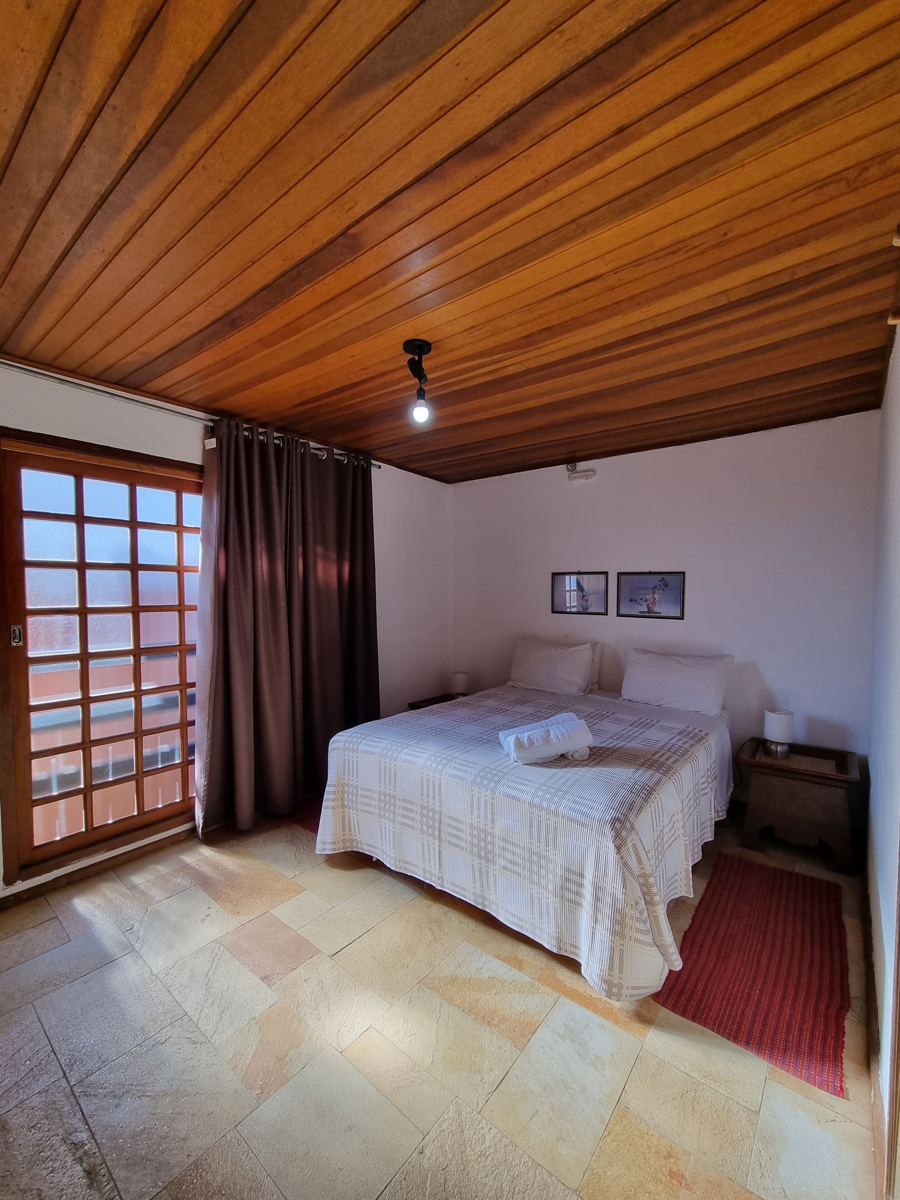 A bedroom with a wooden ceiling, a bed with a patterned bedspread, a nightstand, a red rug, and a window with curtains.