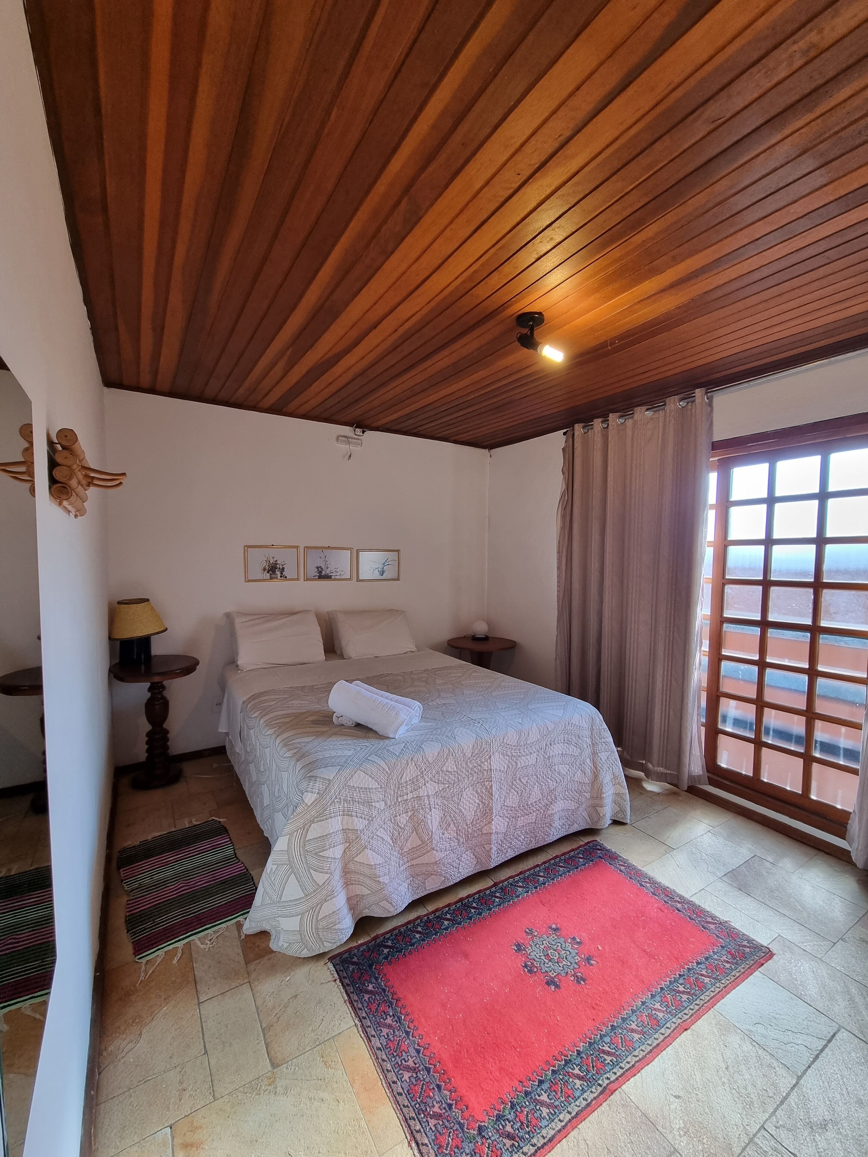 Ein Schlafzimmer mit Holzbalkendecke, einem Bett mit gemustertem Überwurf, einem roten Teppich und Blick durch ein Fenster.