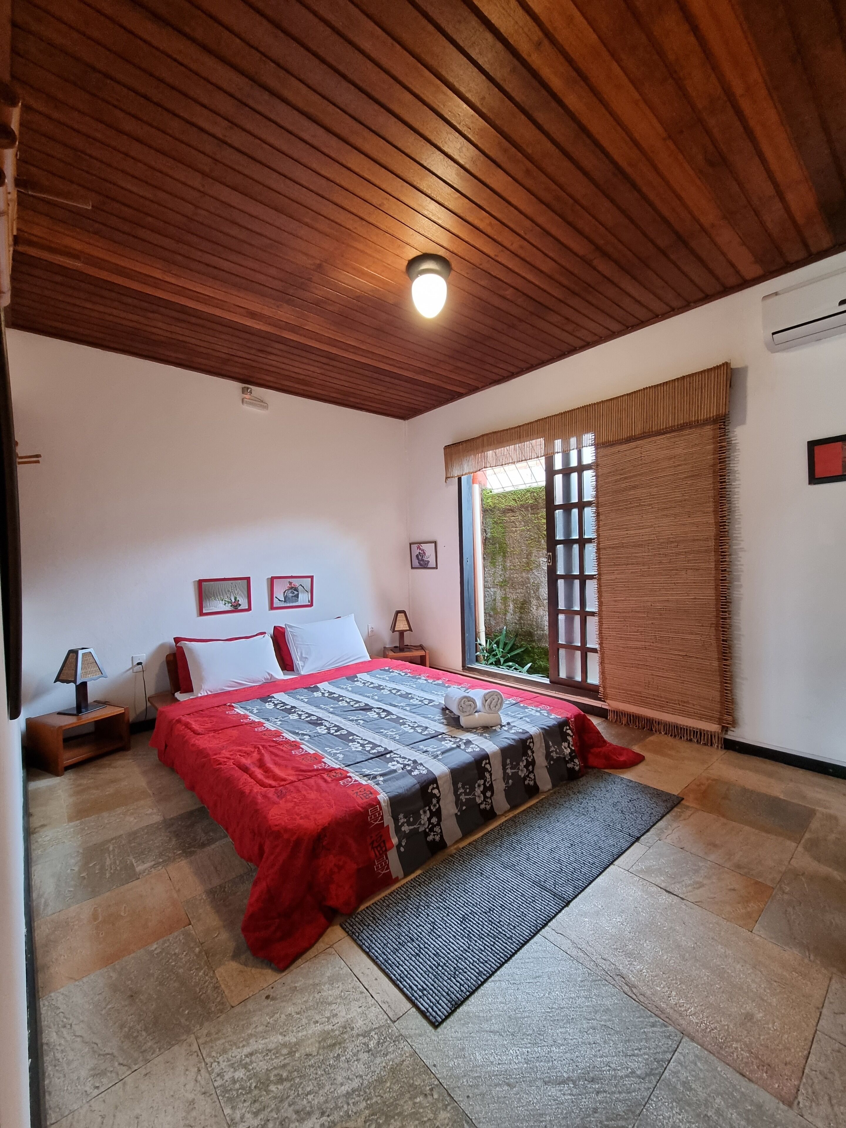 A bedroom with a wooden ceiling, a bed with a red and black bedspread, a nightstand with a lamp, and a window with a view of greenery outside.
