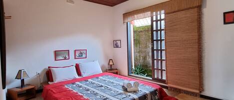 A bedroom with a wooden ceiling, a bed with a red and black bedspread, a nightstand with a lamp, and a window with a view of greenery outside.