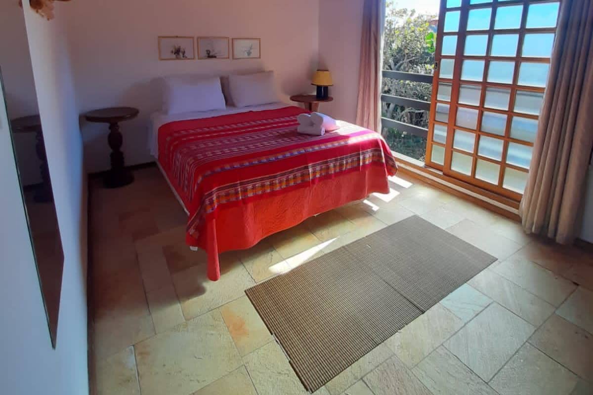A bedroom with a wooden ceiling, a bed with a red bedspread, a small table with a lamp, and a view of the outdoors through a sliding door.