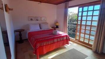 Un dormitorio con techo de madera, una cama con colcha roja, una mesita de noche con lámpara y vistas al exterior a través de una puerta corredera.