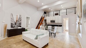 Amplia sala de estar con un sofá blanco, una mesa de comedor con sillas, una escalera y una zona de cocina.