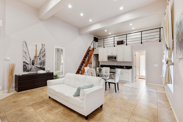 Amplia sala de estar con un sofá blanco, una mesa de comedor con sillas, una escalera y una zona de cocina.