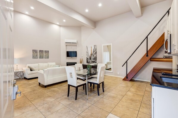 Un vaste espace de vie comprenant un coin repas avec une table, des canapés blancs et une cuisine avec un escalier en bois.
