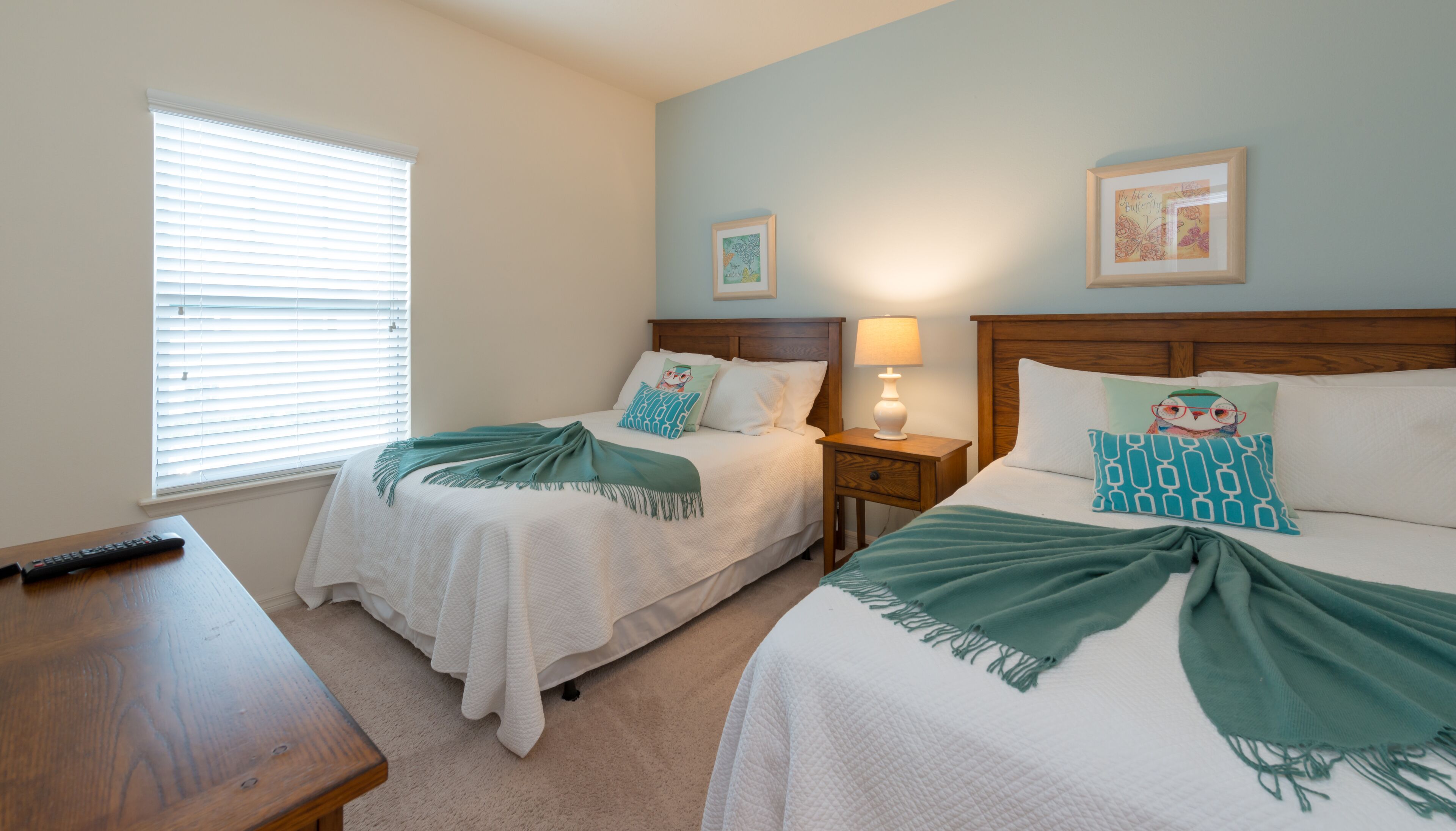 A bedroom with two beds, a bedside table, a lamp, and a window with blinds.