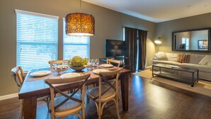 A dining area with a wooden table set for four, a large window with blinds, and a television in the background. - Modern,Stylish & Minutes from Disney (kissimmee)