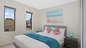 A bedroom with a large bed, two bedside tables, a window with a view of buildings, and a beach painting on the wall. - Astra Apartments Glen Waverley at VIQI (Glen Waverley)