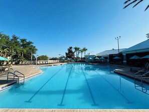 Une piscine d’un bleu éclatant, avec des couloirs de nage, entourée de chaises longues et de parasols, et des palmiers en arrière-plan.