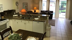 A kitchen with white cabinets, a granite countertop, a sink, and a dishwasher. There is a dining table with chairs, a lamp, and a clock on the wall. - Orlando Oasis (Kissimmee)