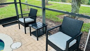 An outdoor patio with two wicker chairs and a small table, a manhole cover, and a view of a grassy area with trees. - Orlando Oasis (Kissimmee)