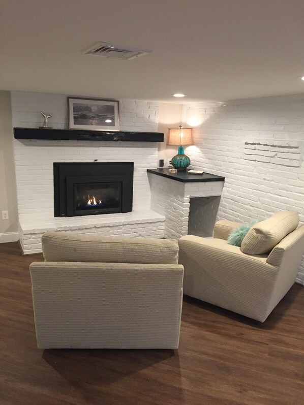 A living room with a fireplace, two white armchairs, a side table with a lamp, and a framed picture on the wall.