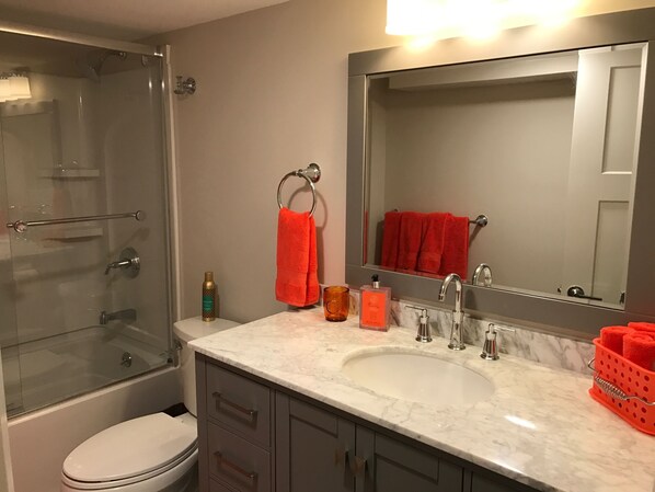 A bathroom with a glass shower enclosure, a white toilet, a sink with a mirror above it, and a towel rack with a red towel.