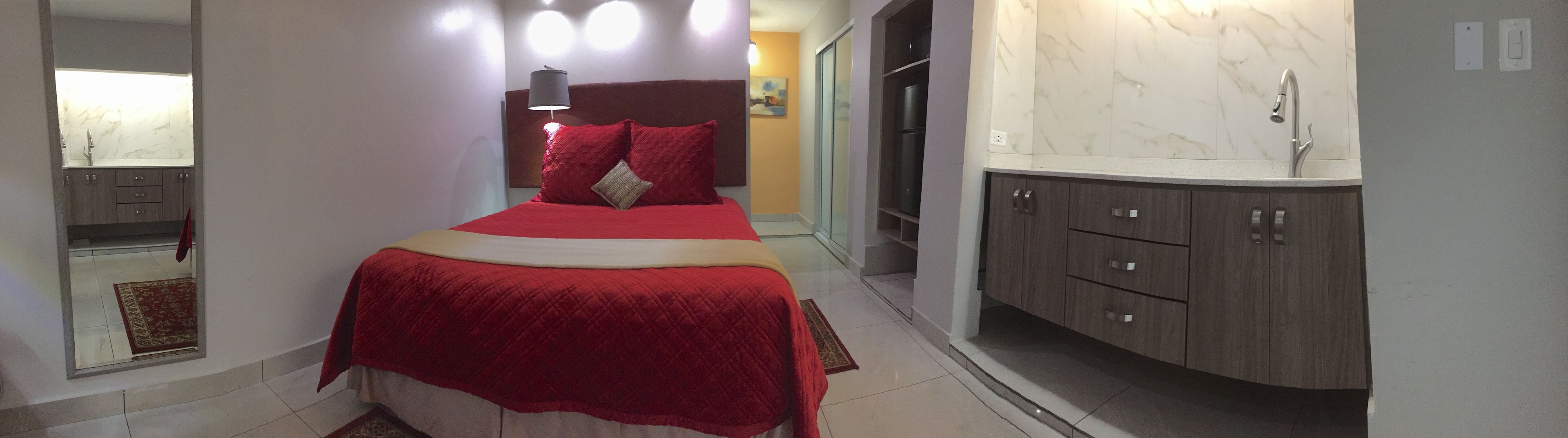 Quarto com cama, mesa de cabeceira, espelho, banheiro com pia e armário, e porta que dá para outro cômodo.