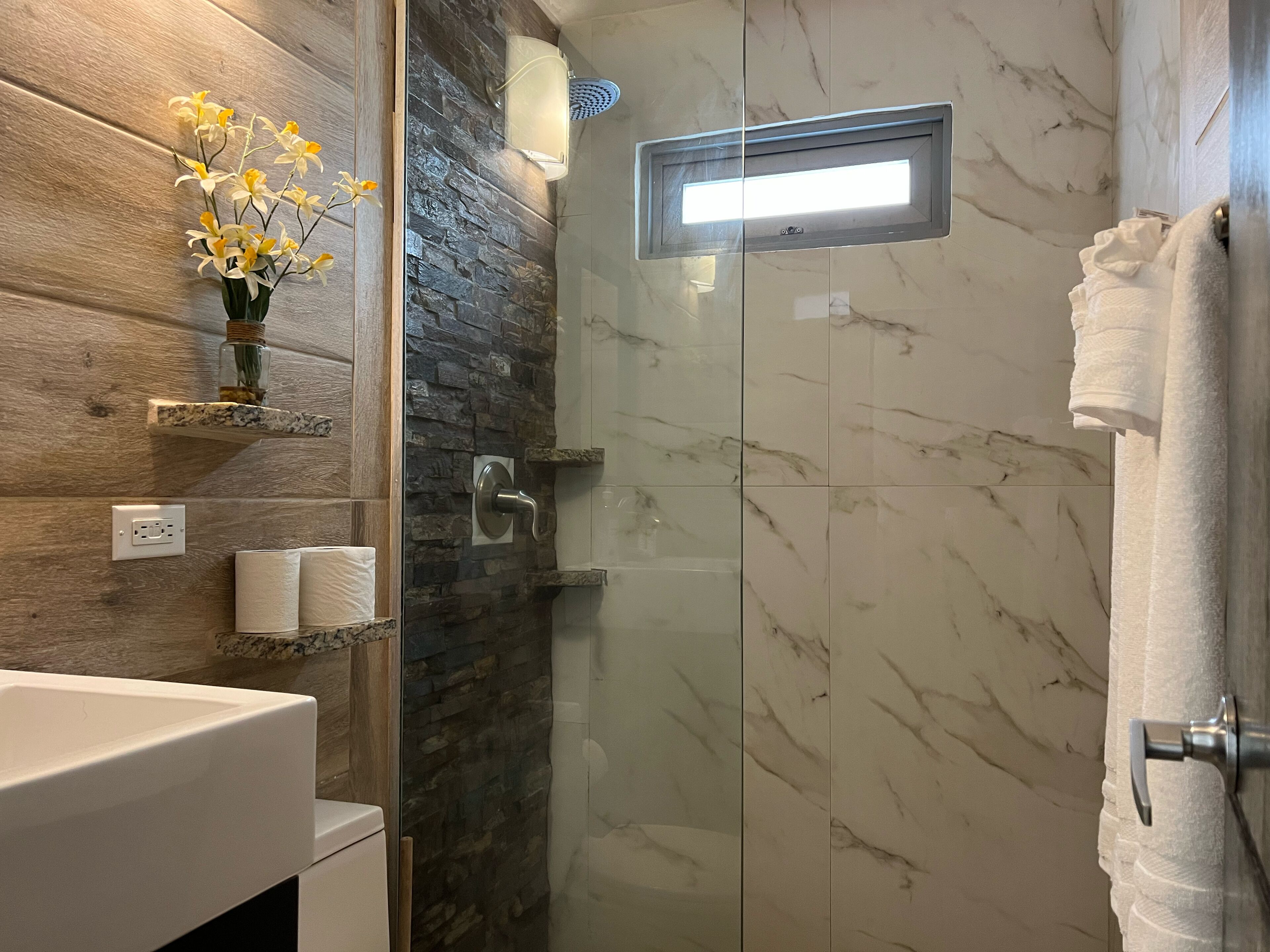 Une salle de bain moderne avec une douche à parois de verre, des carreaux de marbre, un meuble-lavabo en bois et un vase rempli de fleurs jaunes.