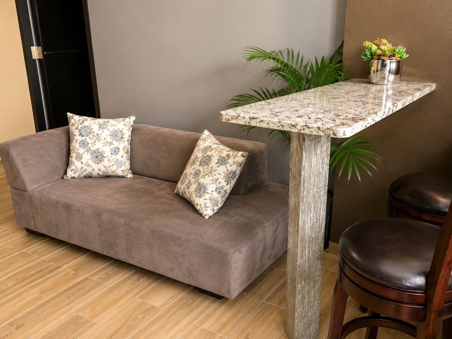 A room with a brown sofa, two patterned pillows, a wooden table, a potted plant, and a leather chair.