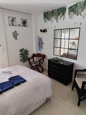 Un dormitorio con cama, cómoda, silla, espejo y decoraciones en la pared, incluyendo plantas y una mandalas.