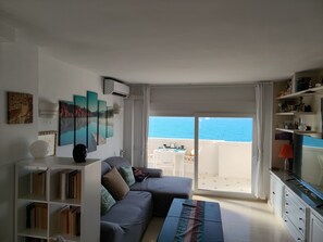 Una sala de estar con un sofá, una estantería, un televisor de pantalla plana y vistas al mar a través de una puerta corredera.